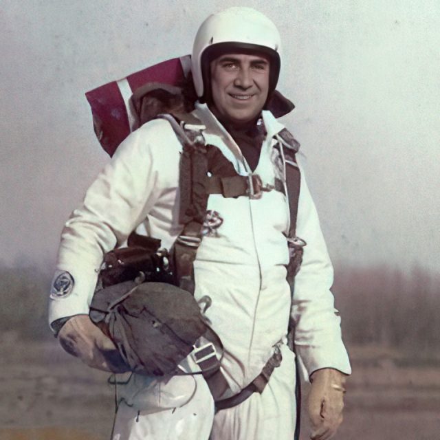 A person wearing a white astronaut suit and helmet stands outdoors, holding a helmet under their arm. The suit has various straps and equipment attached. The background is blurred, giving prominence to the person in the suit.