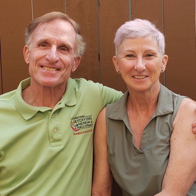 A smiling older couple sits closely together in front of a wooden background. The man wears a green polo shirt, and the woman has short gray hair and wears a sleeveless green top. They're both looking at the camera, radiating warmth and happiness.