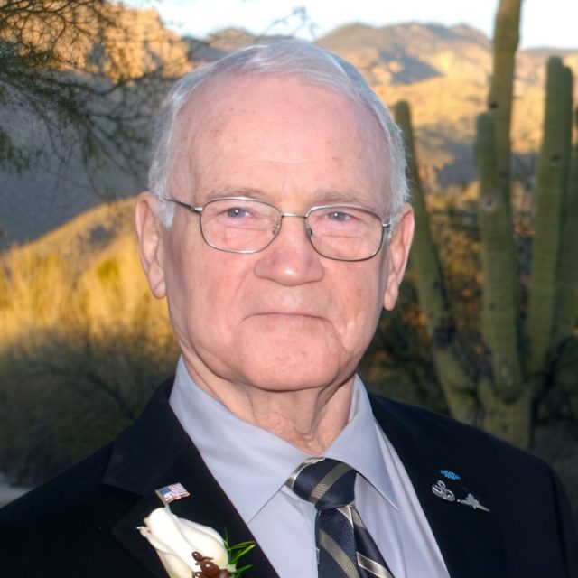 An older man with white hair and glasses stands outdoors against a desert landscape backdrop. He is dressed in a suit with a striped tie and a boutonniere on his lapel. Cacti and mountains are visible in the background.