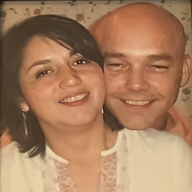 A woman with shoulder-length dark hair and a white blouse smiles next to a bald man wearing an orange shirt. They are in close proximity, appearing warm and happy, with a blurred patterned background.