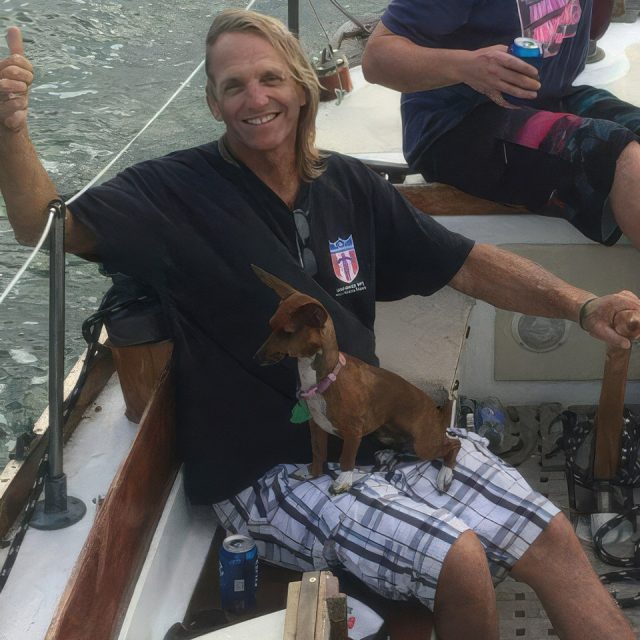 A man wearing a black shirt and plaid shorts sits smiling on a boat, giving a thumbs up. A small brown dog is sitting on his lap. Another person is holding a drink in the background. The boat is on the water.