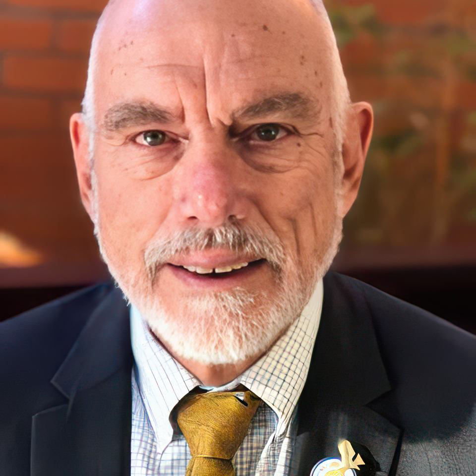 A bald man with a white beard wearing a suit and tie. He is smiling slightly, with a brick wall and greenery in the blurred background.