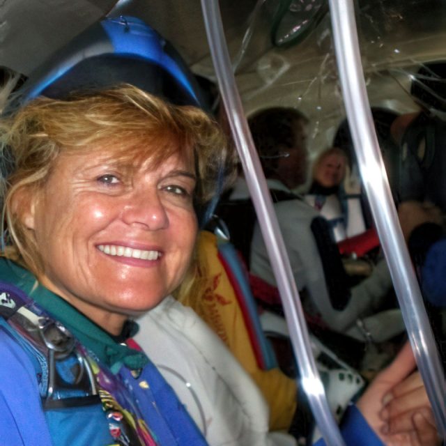 A woman in a blue jumpsuit and helmet smiles at the camera inside an aircraft. In the background, other people sit in skydiving gear. The interior of the plane is visible, suggesting preparation for a skydiving activity.