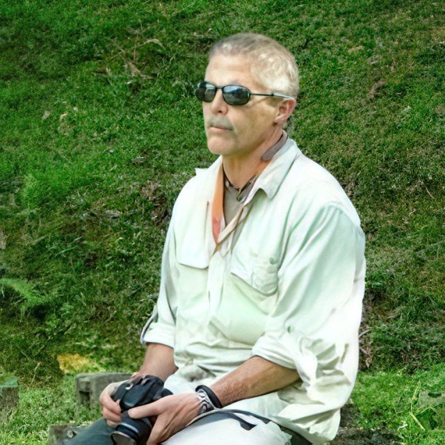 A man in a light shirt and sunglasses sits on a rock outdoors, holding a camera. He is surrounded by green grass and appears contemplative, enjoying a moment in nature.