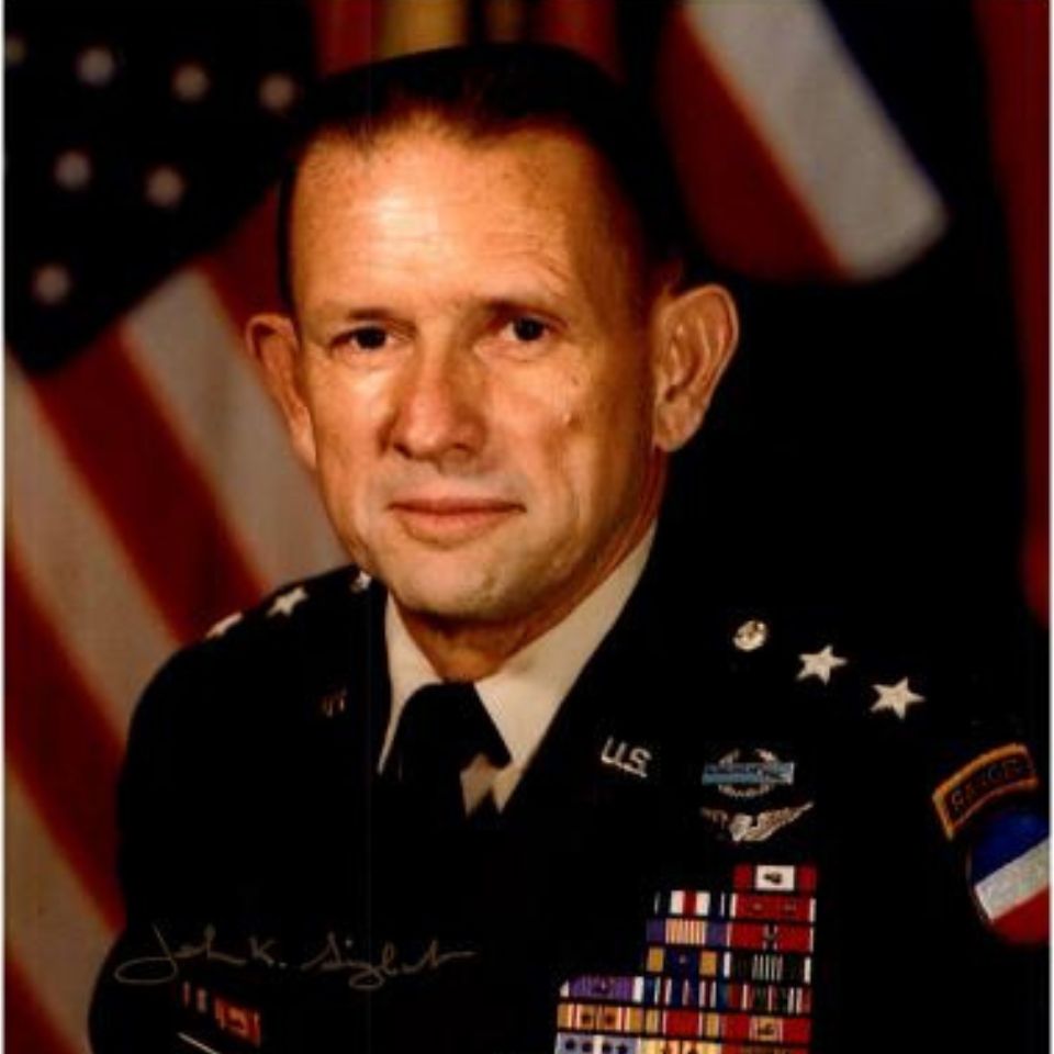 A man in a military uniform with numerous medals and badges poses in front of a U.S. flag. He has short hair and a serious expression.