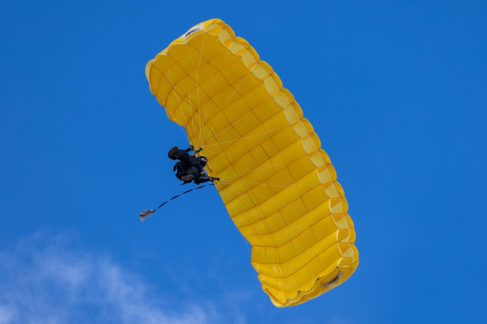 A person is parachuting with a bright yellow parachute against a clear blue sky. The parachute is fully deployed, and the person is wearing dark-colored gear, suspended in the air.