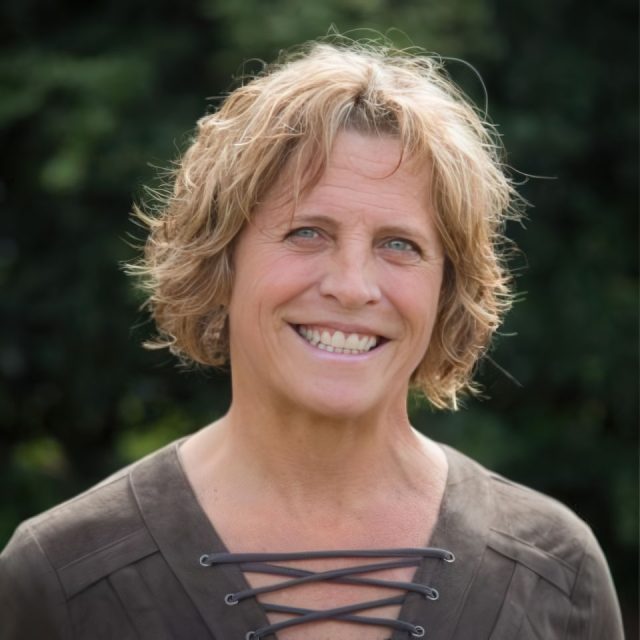 A person with short blond hair and a warm smile stands outdoors. They are wearing a dark, V-neck top with crisscross lacing. The background is a blurred, leafy green.