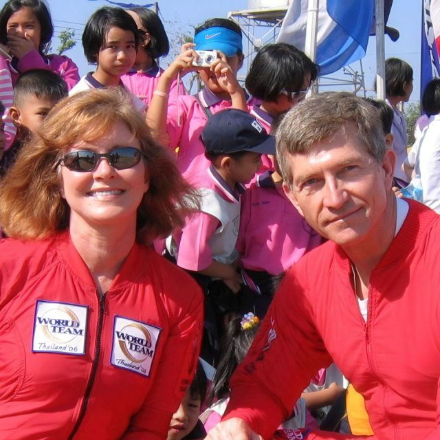 Two people in red jumpsuits with "World Team Thailand 06" patches smile at the camera. They are surrounded by a group of children wearing pink shirts, with some taking pictures. Flags are visible in the background.