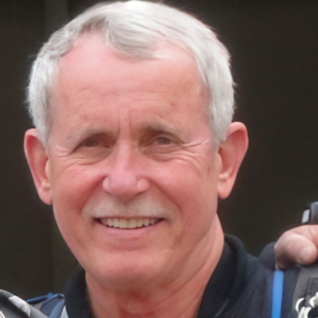 An older man with short gray hair smiles at the camera. He is wearing a dark jacket and appears to be outdoors. The background is blurred.