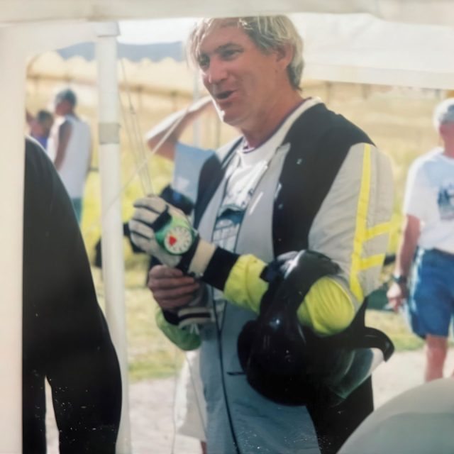 Man in cycling gear holding a helmet and wearing gloves stands under a tent, talking to someone off-frame. Other people are visible in the background on a sunny day.