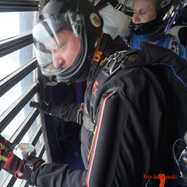 A person wearing a helmet and a skydiving suit looks out of an aircraft window, preparing for a jump. Another person stands behind them, also in skydiving gear. Both appear focused and ready for action.