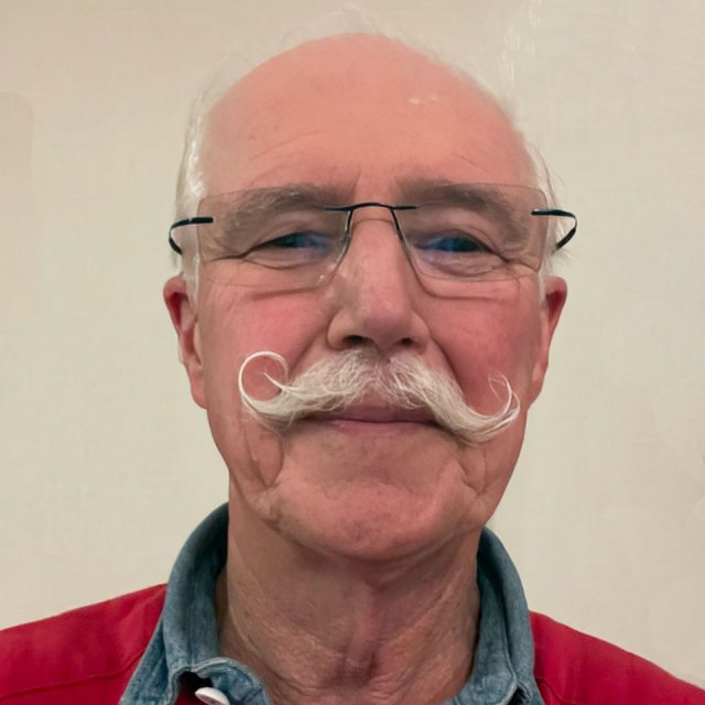 A man with a distinguished curled mustache, wearing glasses. He has a light complexion, a bald head, and is dressed in a red jacket over a denim shirt. The background is a plain, light-colored wall.