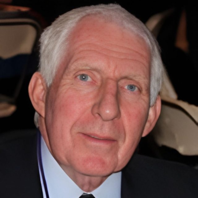 An older man with white hair and blue eyes is seated indoors. He is wearing a suit and tie. Chairs can be seen in the background.