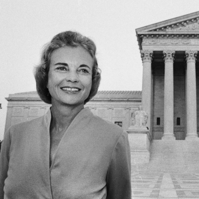 A woman smiles while standing in front of a large, columned building resembling a courthouse. She wears a light-colored jacket, with short hair styled neatly.