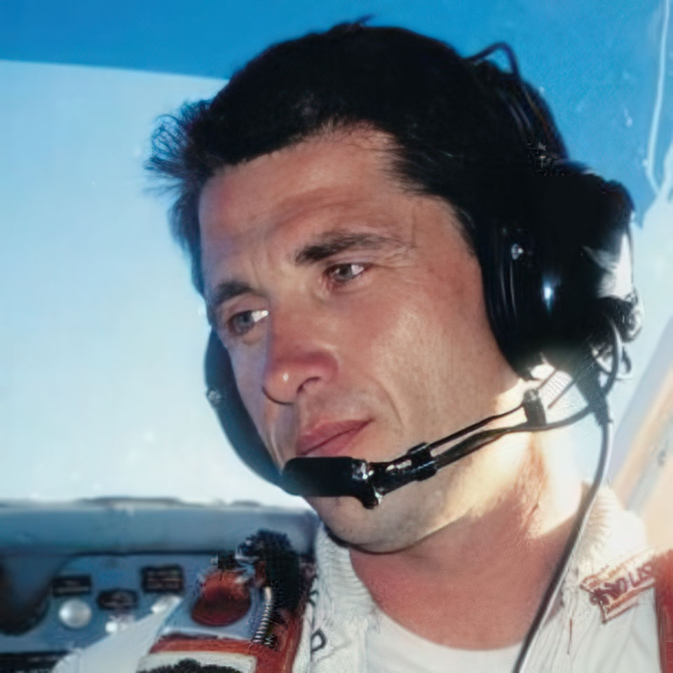 A man wearing a headset and a pilot uniform sits in an aircraft cockpit, looking focused. Sunlight streams through the window, illuminating his face and the controls in front of him.