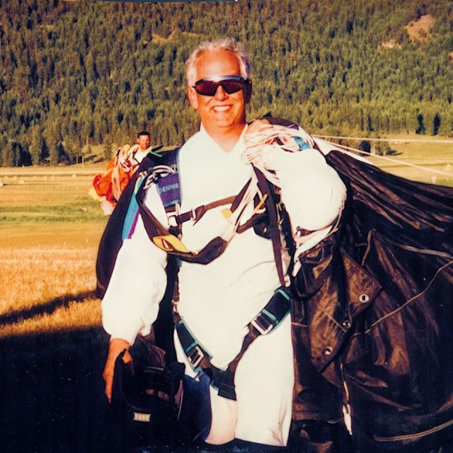 A person in a skydiving suit and harness smiles while holding a parachute. They wear sunglasses and are standing on a grassy field with trees in the background.