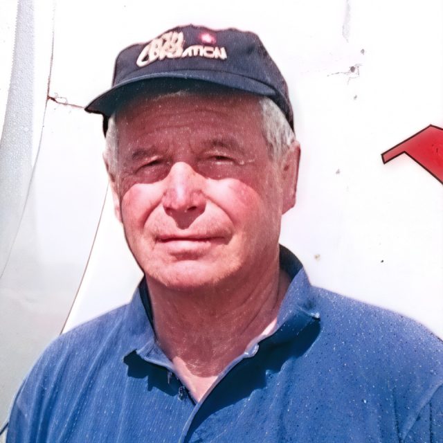 Man in a blue collared shirt and dark cap stands against a white background with partial red lettering visible.