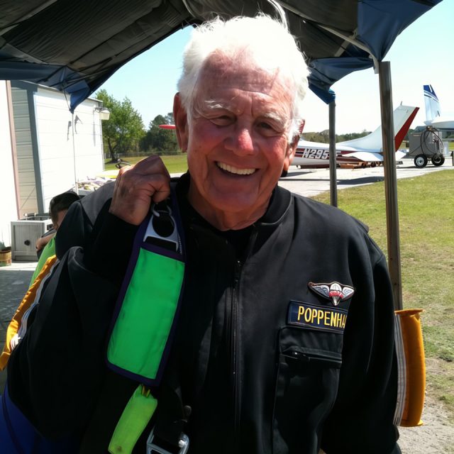 Elderly man with white hair, smiling broadly, wearing a black jacket with a name badge. He's holding a green and blue parachute. Airplanes and a hangar are visible in the background on a sunny day.