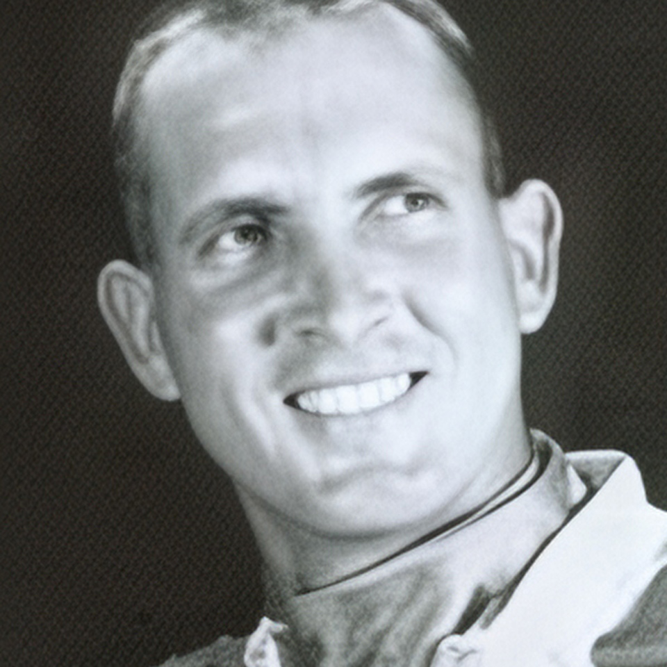 A black and white photograph of a smiling man. He is looking slightly off to the side. The focus is on his face, and he is wearing a light-colored collared shirt. The background is plain and dark.