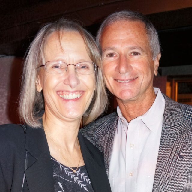 A smiling man and woman stand closely together. The woman has shoulder-length gray hair, glasses, and is wearing a black blazer with a patterned top. The man has short gray hair and is wearing a gray suit jacket with a light-colored shirt.