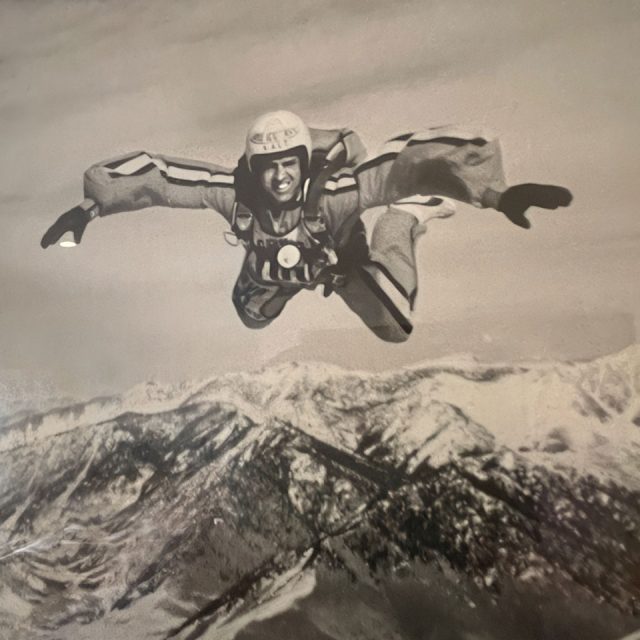 A skydiver in a wingsuit glides above a snow-covered mountain range. The person is smiling and wearing a helmet, captured mid-air against a clear sky.