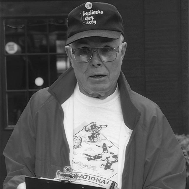 An elderly man wearing a "Skydivers Over Sixty" cap and a skydiving-themed T-shirt stands holding a clipboard. His jacket is open, and he looks directly at the camera. The background is a dark, blurred urban setting.