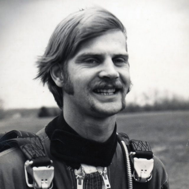 A black and white photo of a man with a mustache and sideburns, smiling. He's wearing a parachute harness and standing outdoors, with a blurred landscape in the background.