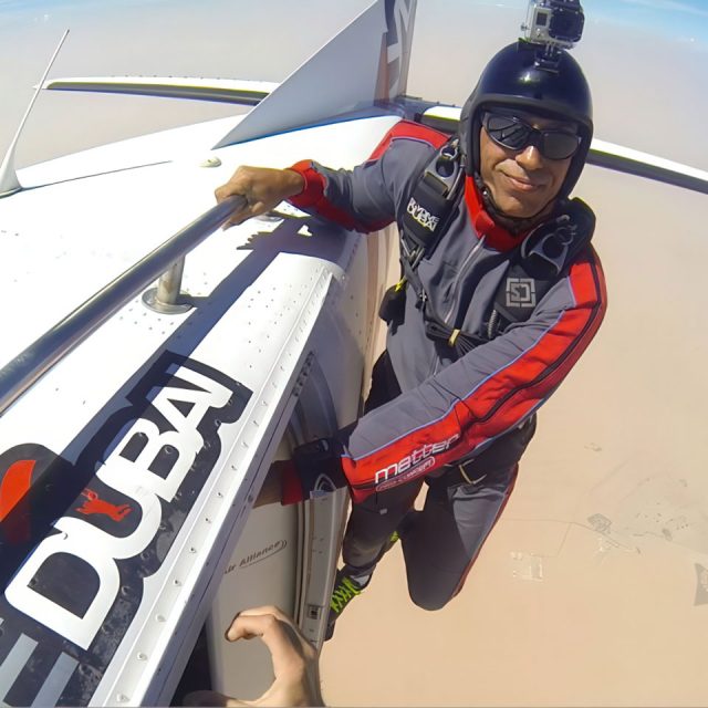 Skydiver in a jumpsuit with a GoPro camera mounted on their helmet stands at the open door of a plane high above desert terrain. The plane has a "Dubai" logo on the side.