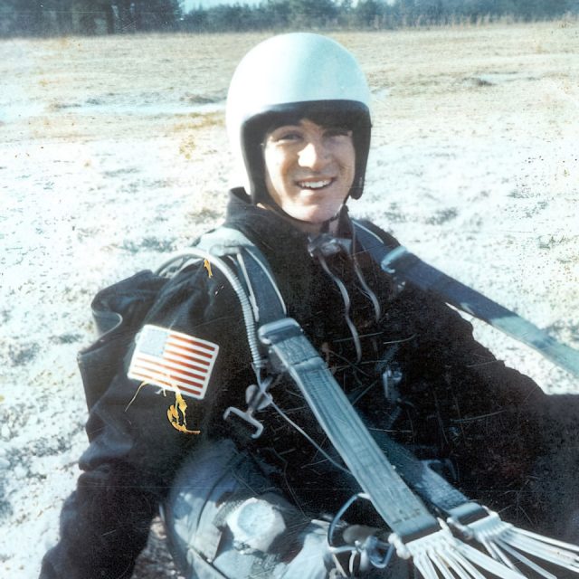 A person in a skydiving suit and helmet sits on the ground with parachute straps visible. They are outdoors on a sandy surface, smiling, with an American flag patch on their sleeve.