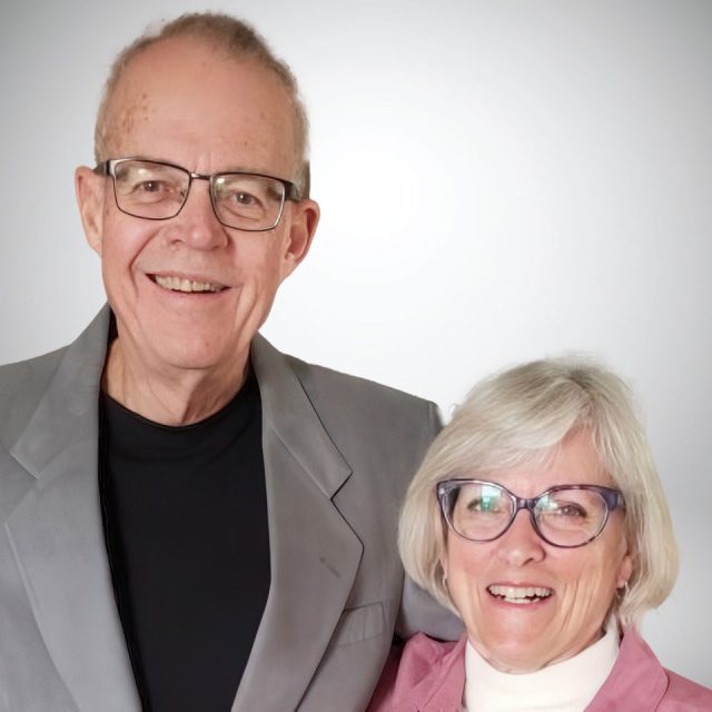 An older couple standing together smiling. The man wears glasses, a gray blazer, and a black shirt. The woman also wears glasses, a white top, and a pink blazer. The background is plain and light-colored.