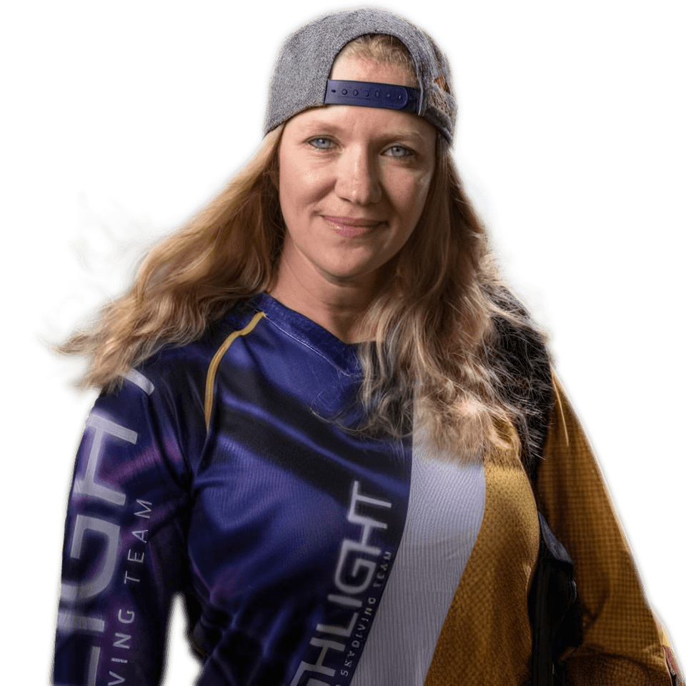 A woman with long blonde hair wearing a grey cap and a colorful jersey stands facing the camera. She has a subtle smile and a relaxed demeanor. The jersey has text on it, but it’s not fully clear. The background is plain.