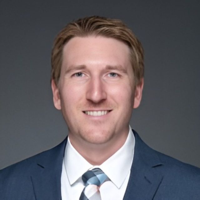 A person with short, light brown hair is smiling, wearing a dark suit, white shirt, and striped tie, against a gray background.