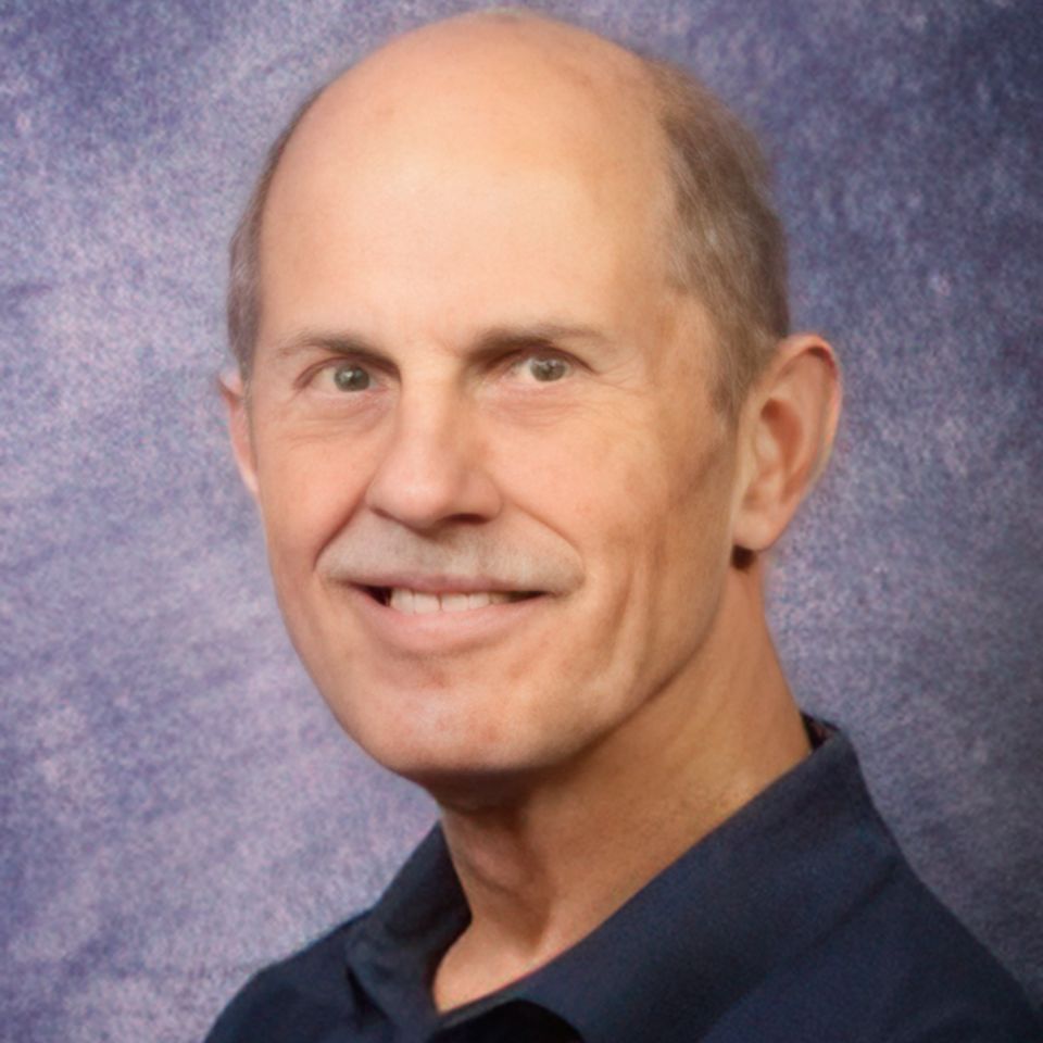 A middle-aged man with a bald head and a friendly smile is wearing a dark blue shirt. The background is a mottled grayish-purple.