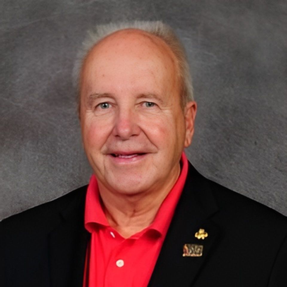 A smiling man with short, light-colored hair wearing a bright red shirt and a black blazer, featuring lapel pins. The background is a plain, textured gray.