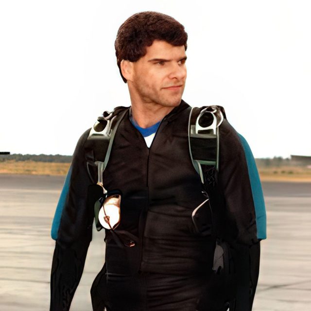 A person in a black parachuting suit stands on an airfield with a confident expression, gazing to the side. They are wearing a parachute harness and the background shows a runway and clear sky.