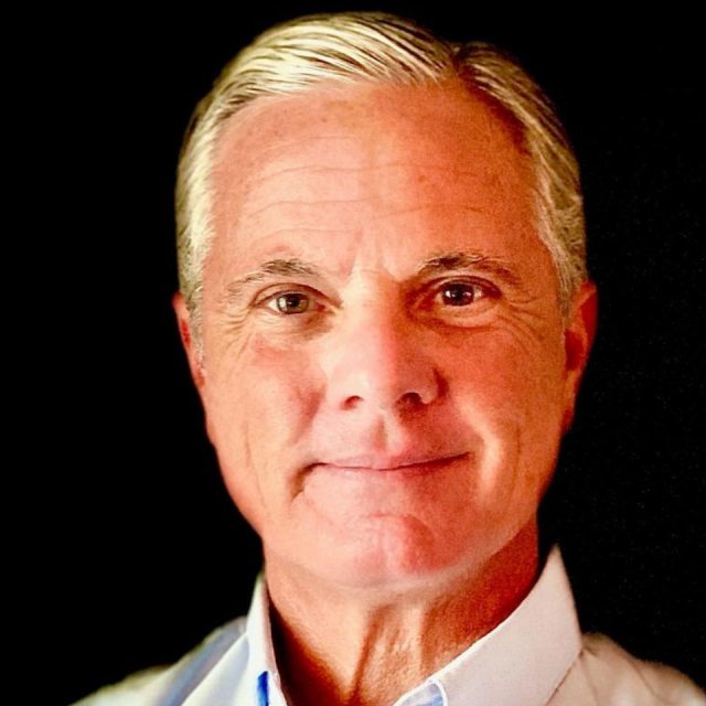 Portrait of a smiling man with short gray hair, wearing a light-colored shirt, set against a black background.