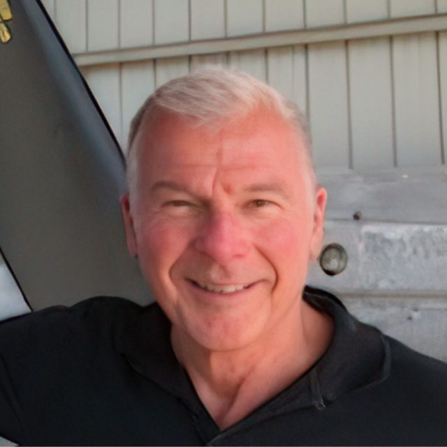 A smiling man with short white hair, wearing a black shirt, stands in front of a gray industrial background.