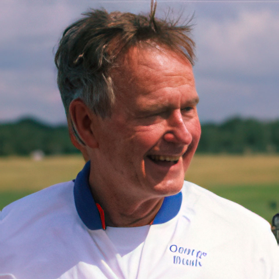 A smiling older man in a white polo shirt, standing outside with a blurred grassy landscape in the background. His hair is slightly tousled, and he appears relaxed and content under a partly cloudy sky.