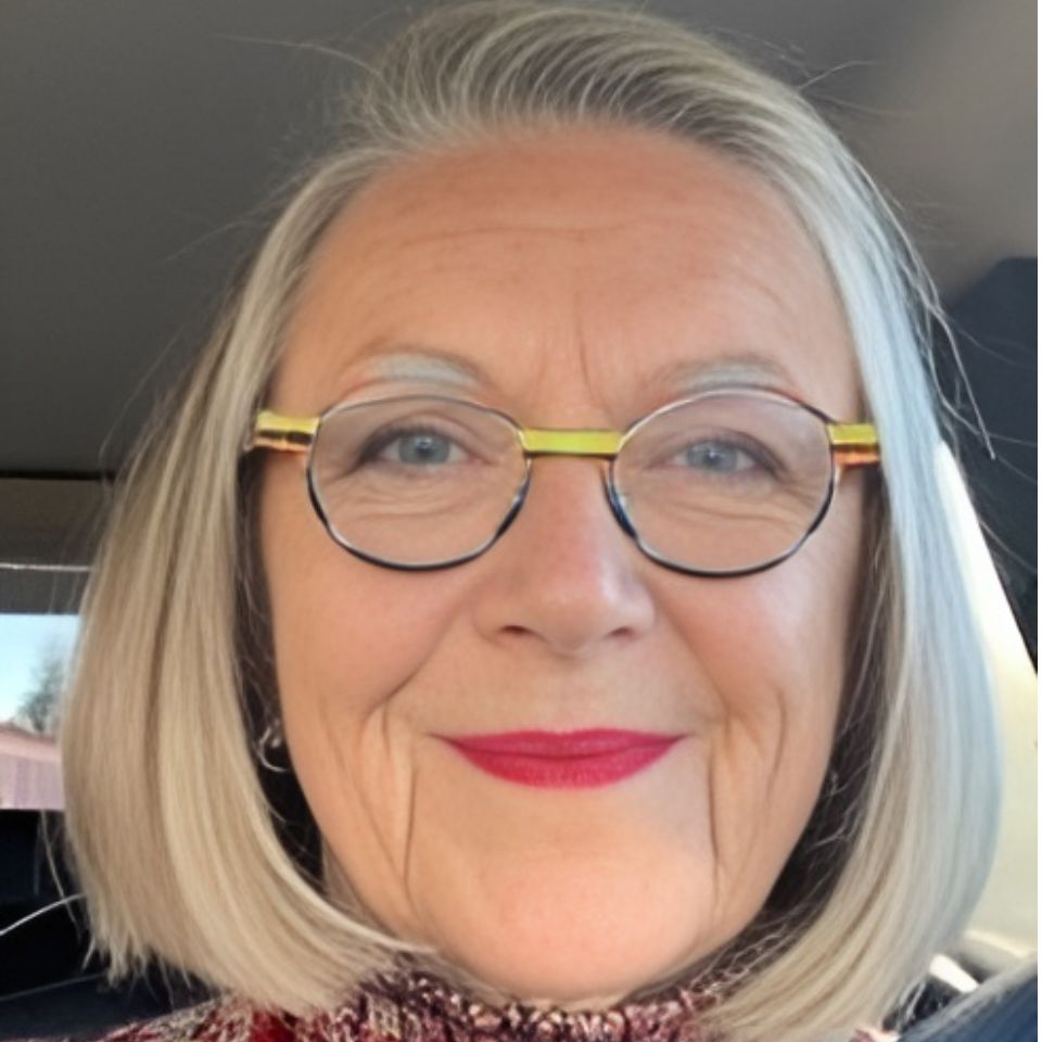 A woman with short gray hair and round glasses is smiling warmly. She wears red lipstick and a patterned top, and is sitting inside a car. The background is slightly blurred.