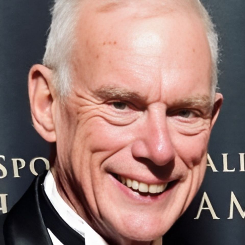 Smiling older man in a tuxedo, standing against a dark background with text partially visible.