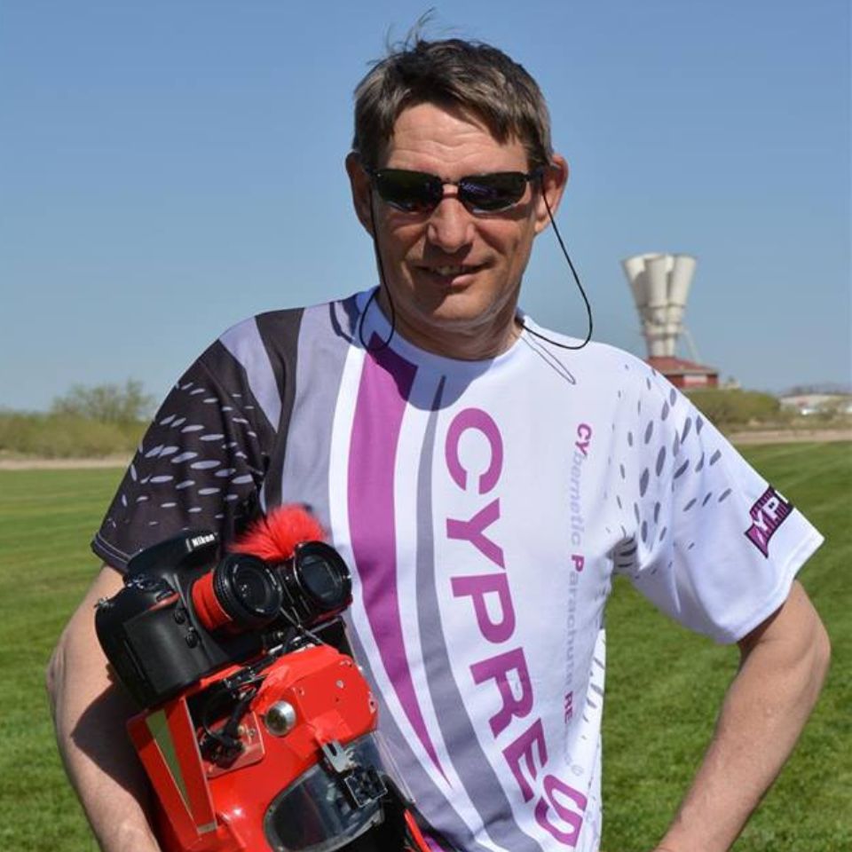 A person wearing sunglasses and a "CYPRES" t-shirt stands outdoors on a grassy field, holding a red camera with accessories. The sky is clear, and a distinctive white structure is visible in the background.