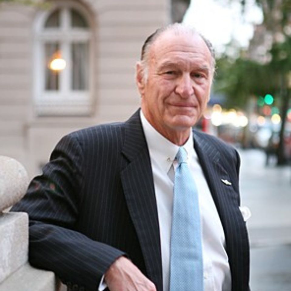 Elderly man in a suit and light blue tie leans against a stone railing in an urban setting. A building and trees are in the background. The scene is well lit and appears to be taken during daytime.