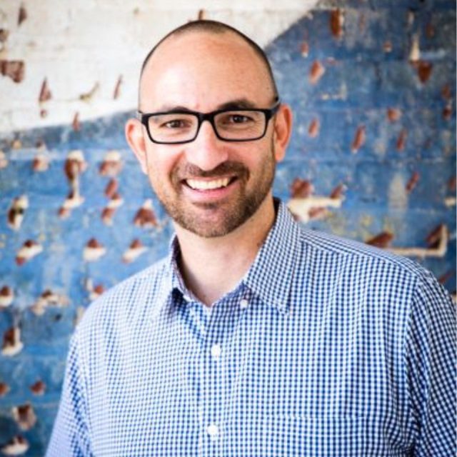 A smiling man with glasses and a beard stands in front of a textured wall. He is wearing a checkered shirt, and the background features a blue and white rough painted pattern.