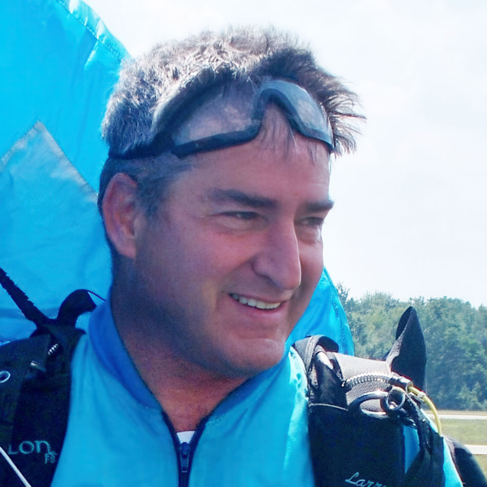 A man in a blue skydiving suit smiles, wearing a protective goggle on his forehead. A parachute is visible in the background against a clear sky and distant trees.