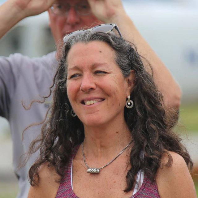 A woman with long, curly hair and sunglasses on her head smiles at the camera. She is wearing a necklace, earrings, and a sleeveless top. A man in the background adjusts his glasses. The setting appears outdoors.