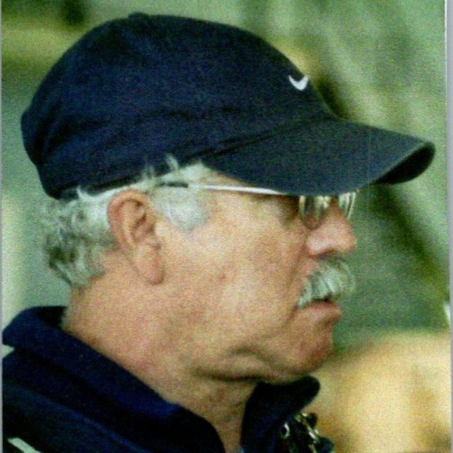 An elderly man with gray hair and a mustache is wearing glasses, a navy blue cap, and a matching jacket. He is looking to the side, appearing focused.