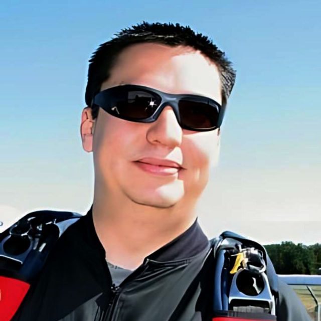 A person wearing sunglasses and a skydiving suit smiles outdoors on a clear day. The harness and gear suggest readiness for skydiving. The background shows a blue sky and some distant trees.