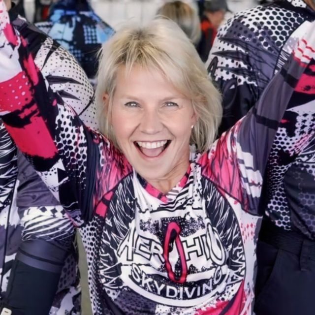 A smiling skydiver raises her arms in excitement, wearing a colorful jumpsuit that reads "Skydiving" in bold letters. The background shows several others in similar attire, suggesting a group event or gathering.