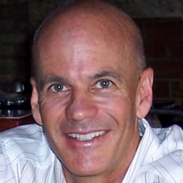 A smiling man with a shaved head wearing a white, striped shirt is looking at the camera. The background is dark and slightly blurred.