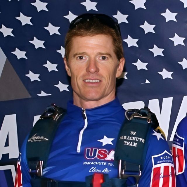 A person wearing a blue USA Parachute Team jumpsuit with harnesses stands in front of a backdrop featuring white stars on a dark background. The individual is smiling and has short, reddish-brown hair.